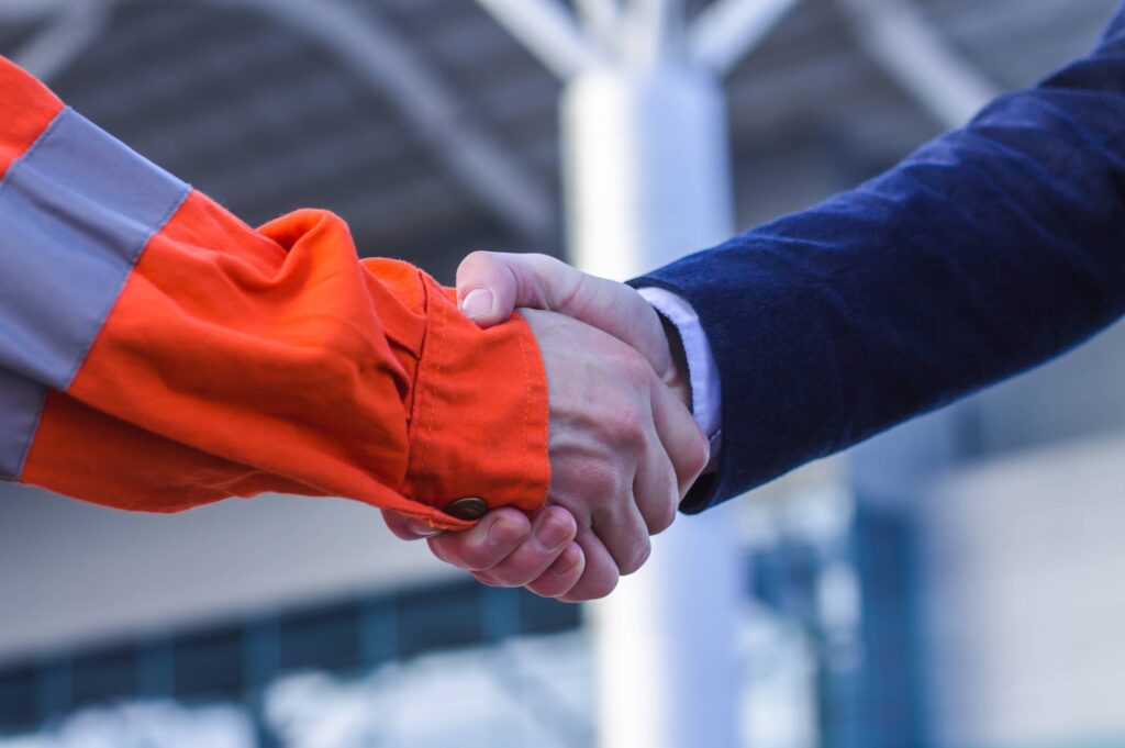 A close up phone of a handshake between an individual wearing bright orange protective jacket and someone wearing a blue suit. The background is obscured, though appears to be in front of a building.