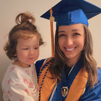 A smiling young woman in a graduation cap holding a toddler in her arms
