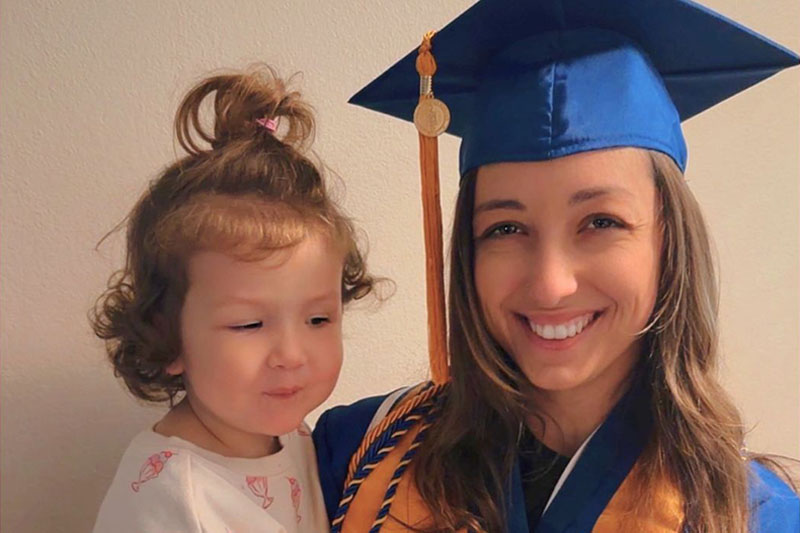 A smiling young woman in a graduation cap holding a toddler in her arms 