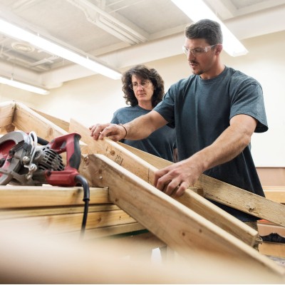 two people working with raw lumber, power tool in the foreground